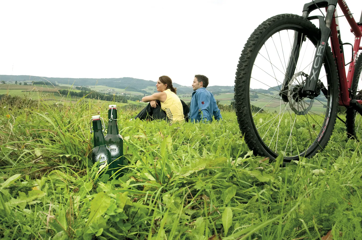 Zwei Freunde sitzen mit Bier in einer Wiese neben ihren Fahrrädern und blicken in die Landschaft