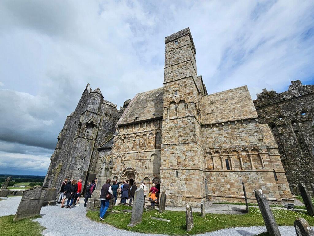 Seitenansicht des Rock of Cashel mit dem St. Patrick's Cross im Vordergrund und den historischen Gebäuden der Anlage im Hintergrund.
