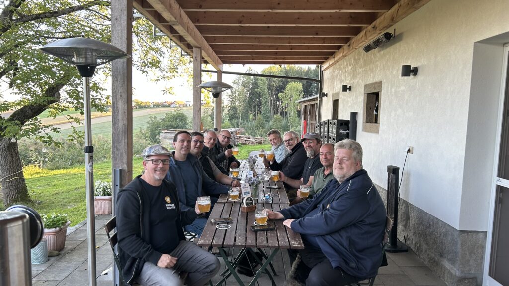 Die Gruppe mit Biergläsern an einer langen Holztafel auf der überdachten Terrasse in Kronedt, dahinter Wiesen und Bäume.