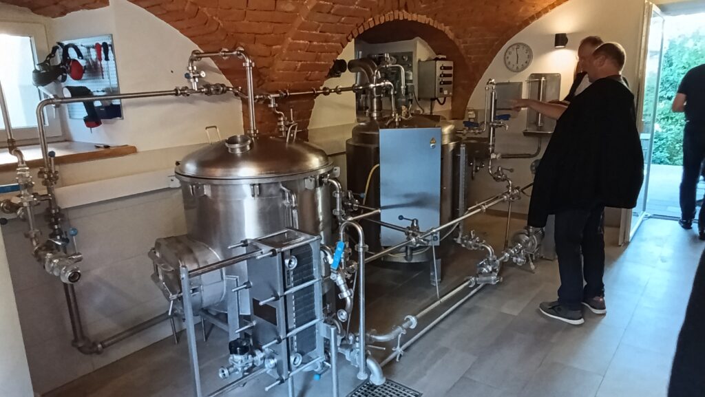 Kleine Brauanlage mit Edelstahlkesseln und Rohrleitungen unter einem Ziegelgewölbe, daneben Besucher bei der Besichtigung.