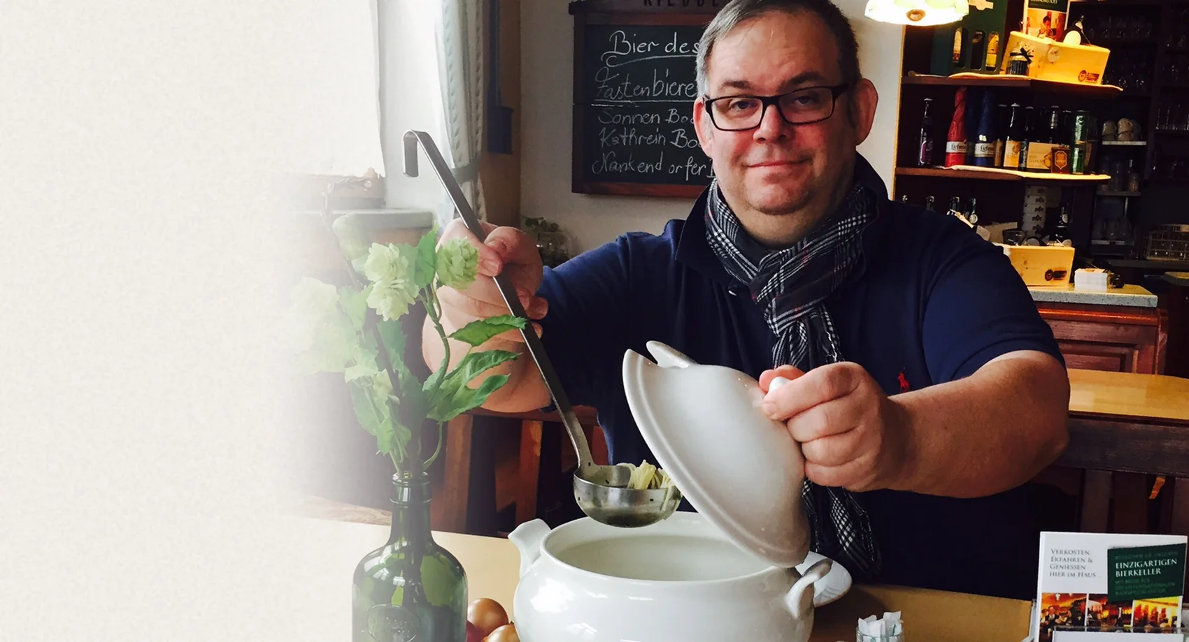 Karl Zuser jun. schöpft mit dem Suppenschöpfer Buchstabensuppe aus einer weißen Suppenterrine, daneben Hopfen in einer Bierflasche, im Hintergrund die Tafel „Bier des Monats“ im Biergasthof Riedberg
